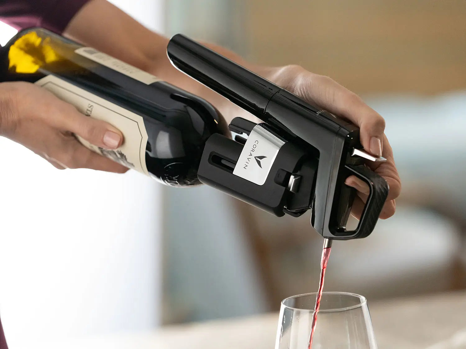 A hand using a Coravin wine opener to pour red wine from a bottle into a glass.