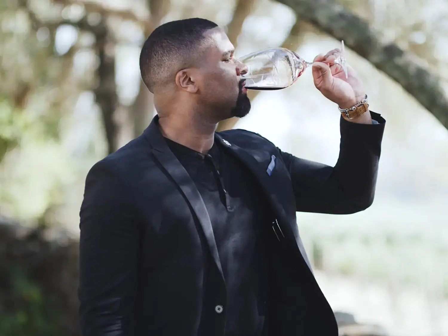 A man in a black blazer sipping red wine from a glass outdoors, surrounded by trees and natural greenery.
