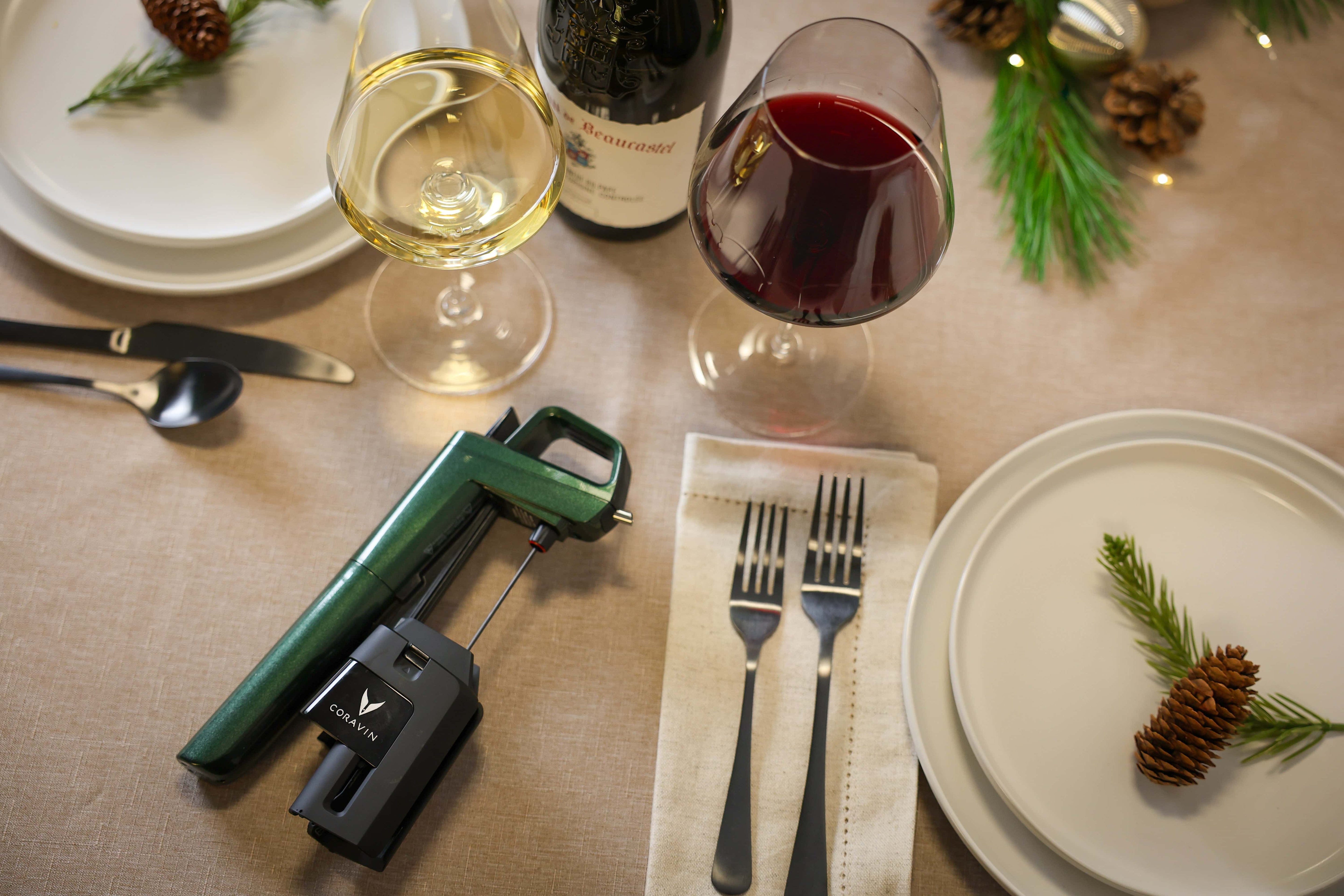 A dining table setting with two wine glasses, a Coravin wine opener, plates, forks, pinecones, and greenery accents.