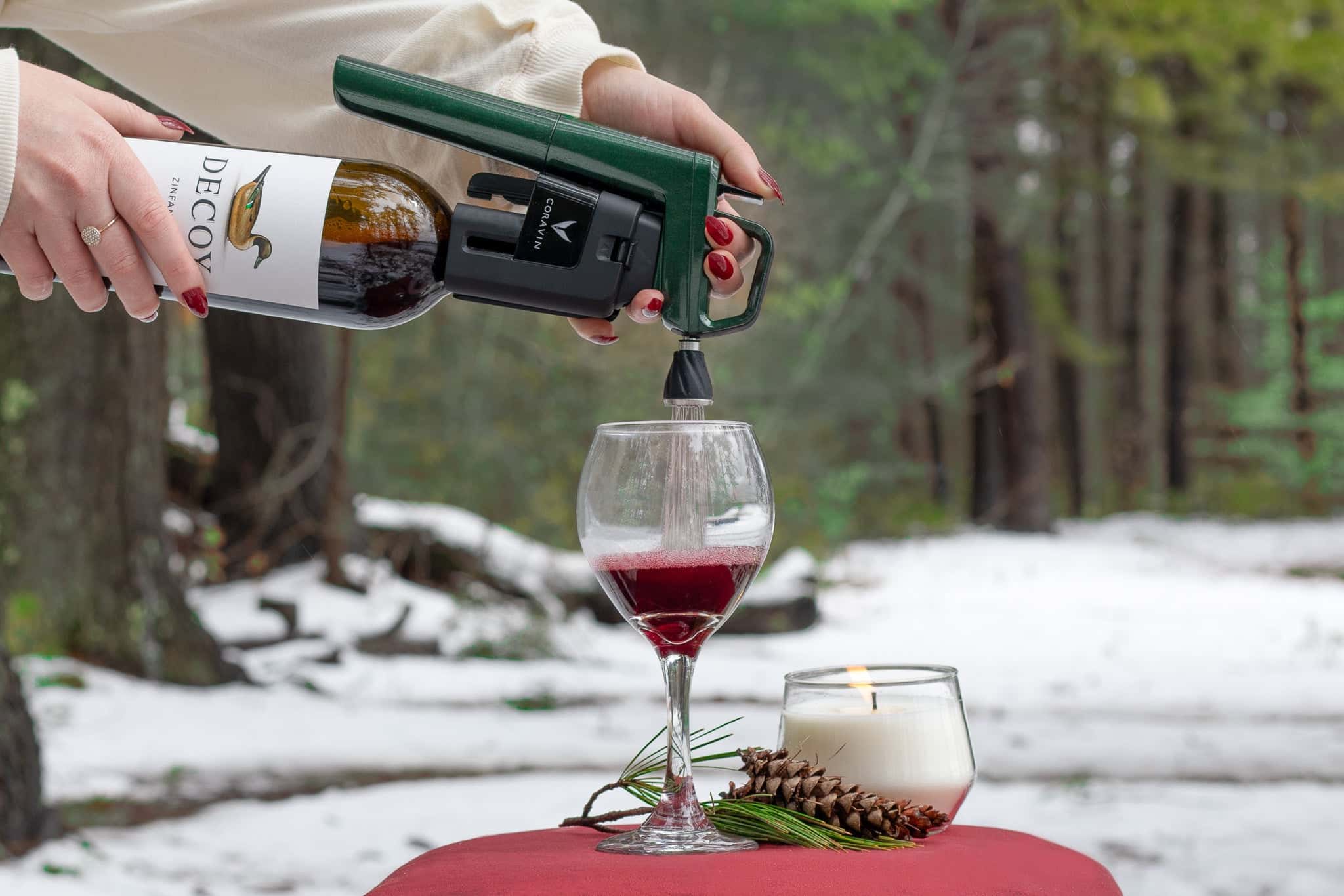 A woman pours wine from a Coravin into a glass, with a lit candle and pinecone on a red table, surrounded by snow.
