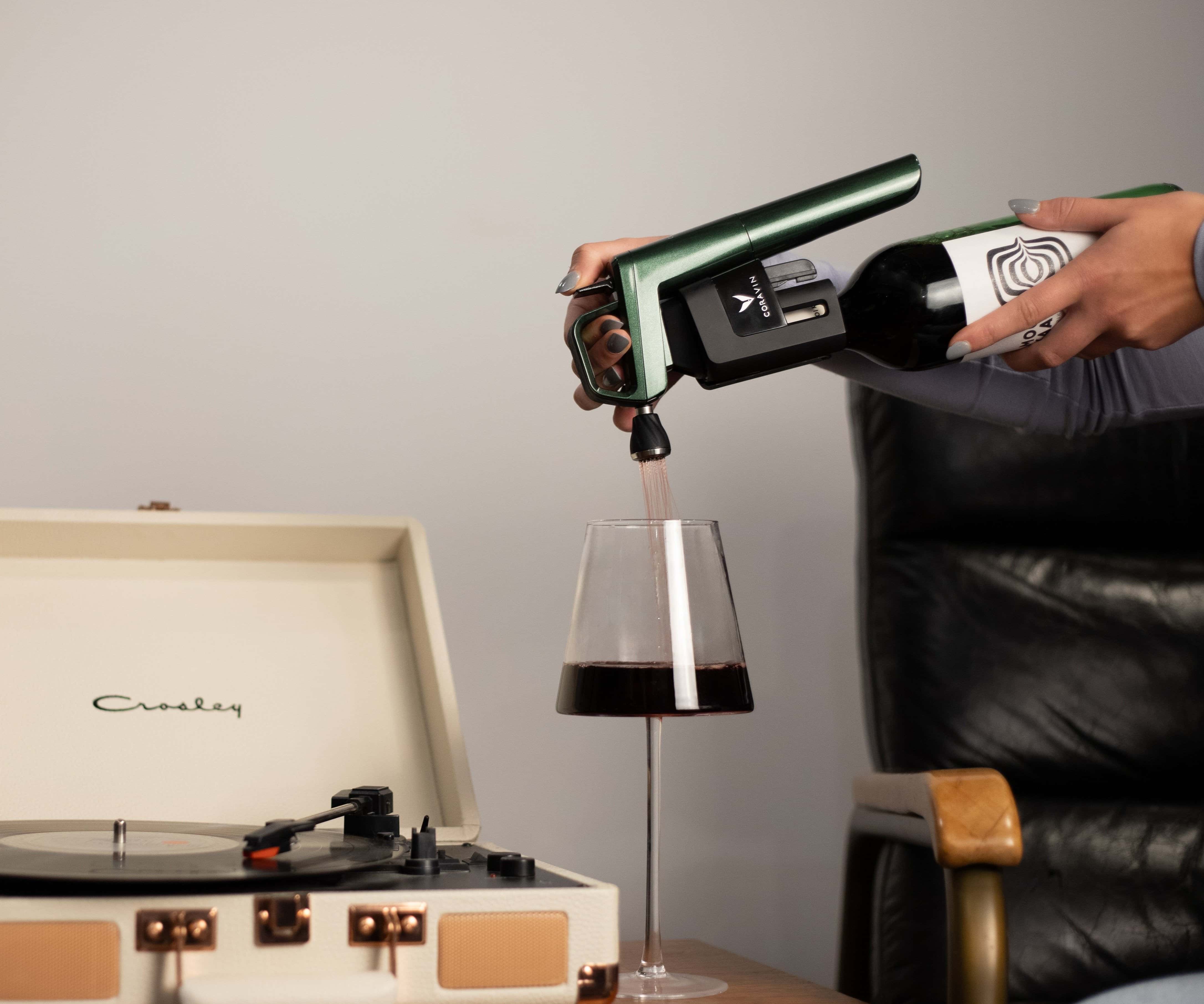 A hand using a green wine aerator to pour red wine into a glass beside a vintage record player.