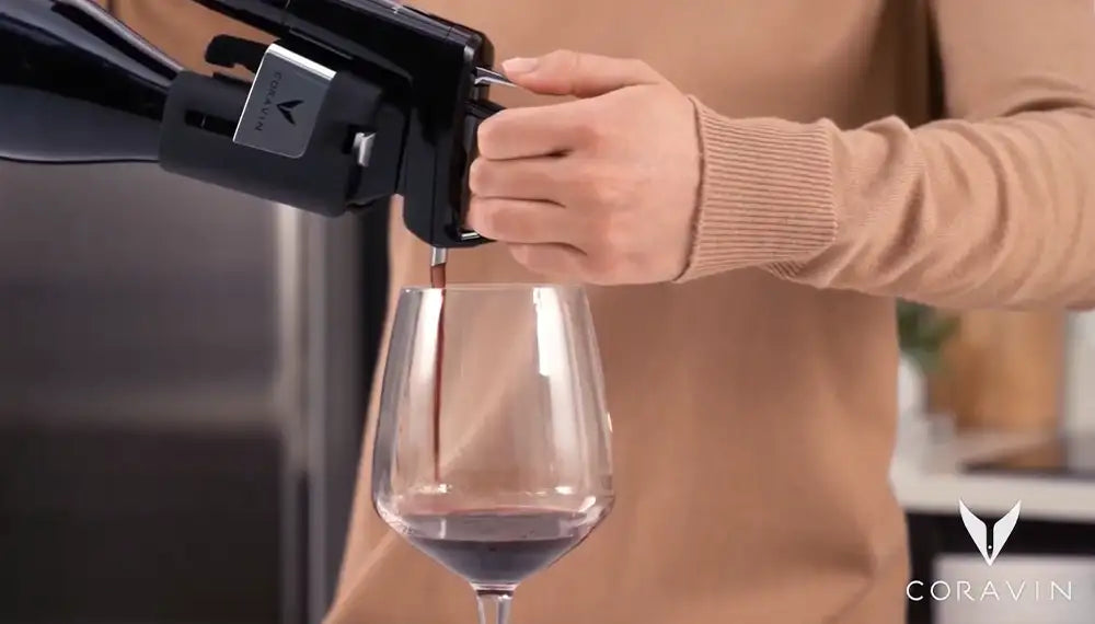 A hand using a Coravin wine preservation system to pour red wine into a glass.