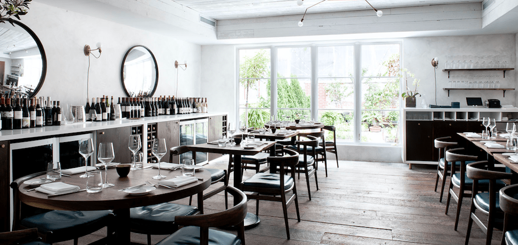 Elegant dining room at The Musket Room with natural light, wooden floors, minimalist decor, and a curated wall of fine wine bottles.