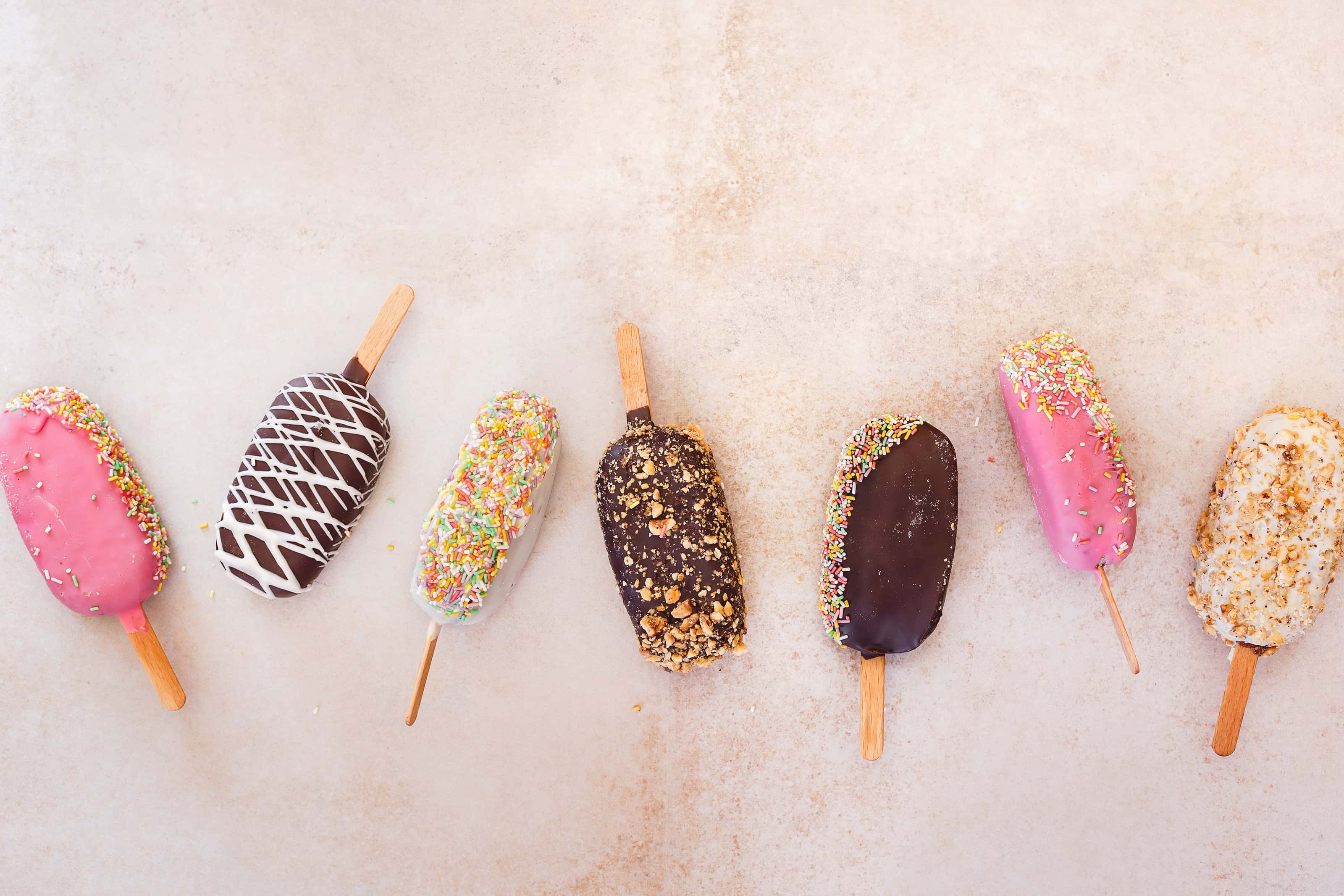 Assorted colorful ice cream bars on wooden sticks coated with chocolate, pink glaze, sprinkles, and crushed nuts, displayed on a light beige background.