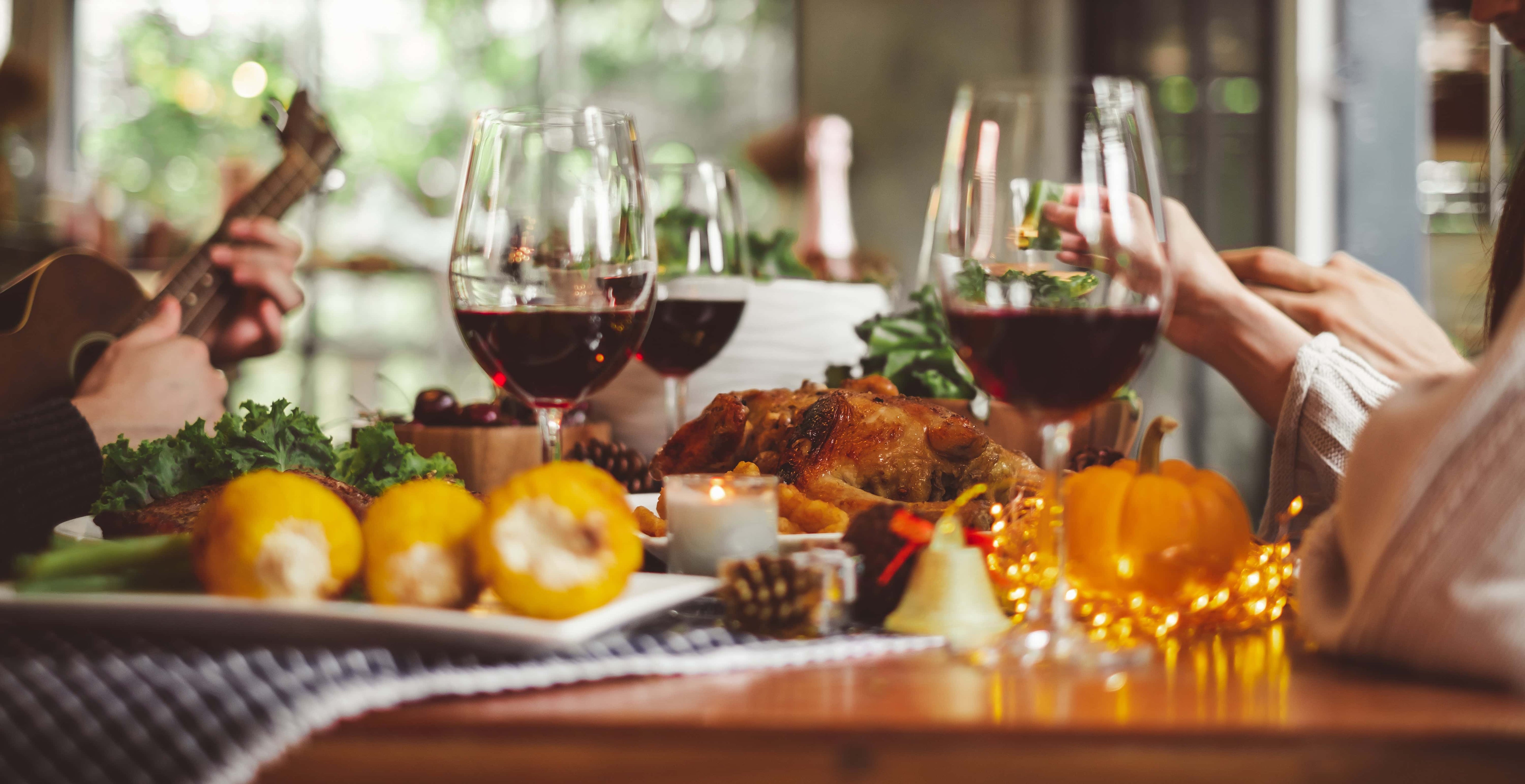 Warm Thanksgiving or fall gathering scene with a table set for dinner, featuring roast turkey, corn, wine glasses filled with red wine, and decorative pumpkins, while people dine and one person plays a ukulele.