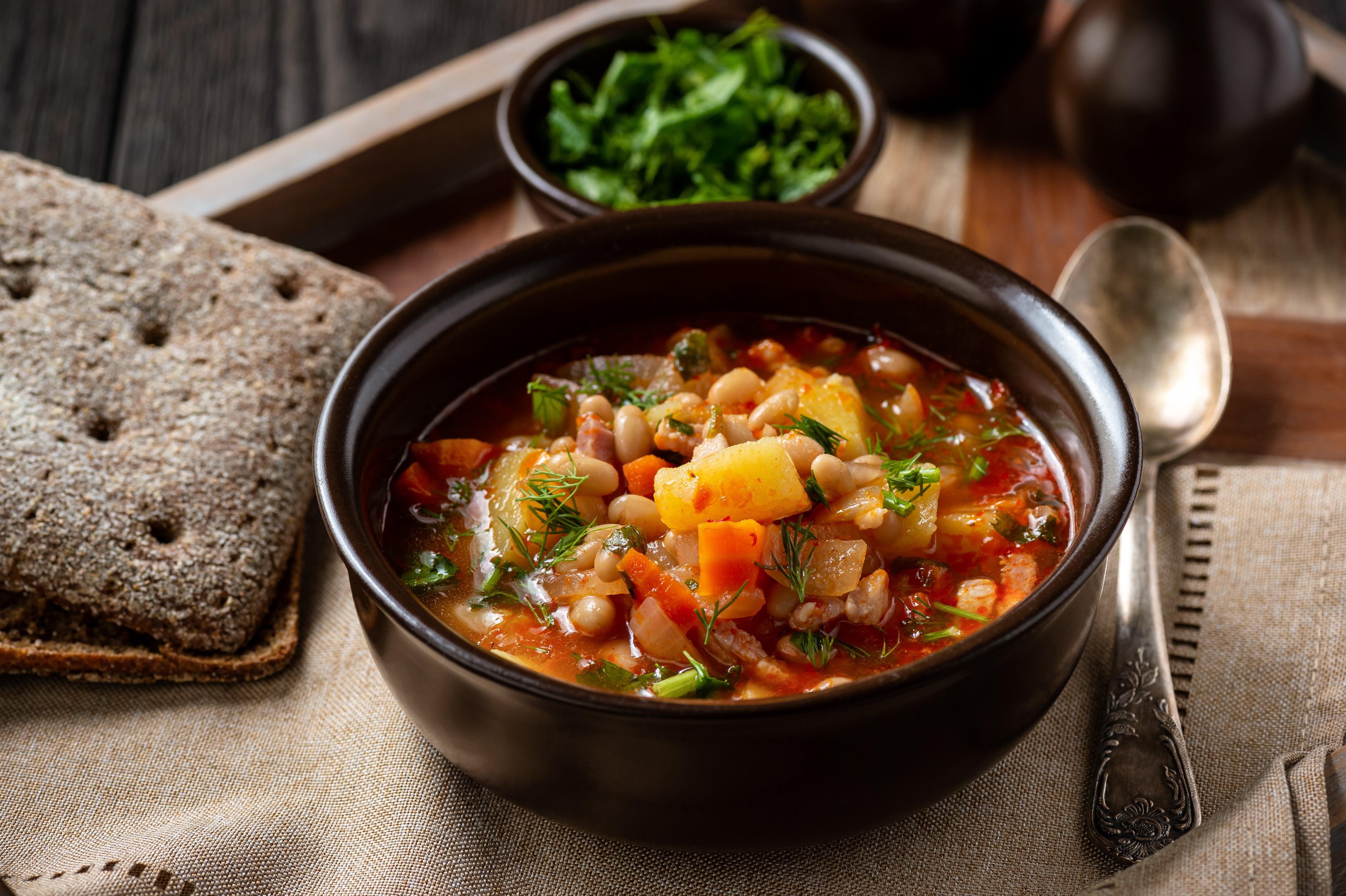Hearty bowl of vegetable and bean soup garnished with fresh herbs, served with crisp rye bread and a side of chopped greens.