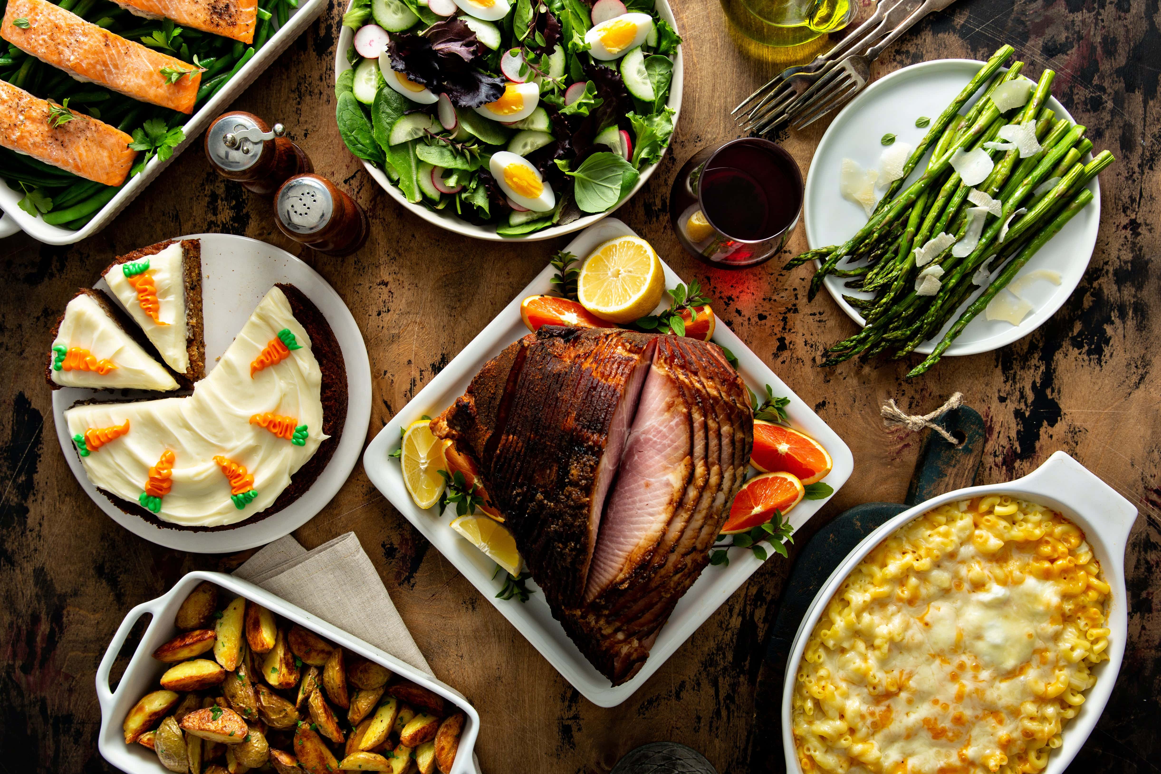 Festive dinner table spread featuring glazed ham, roasted potatoes, baked salmon, macaroni and cheese, asparagus with parmesan, fresh salad, and carrot cake dessert.