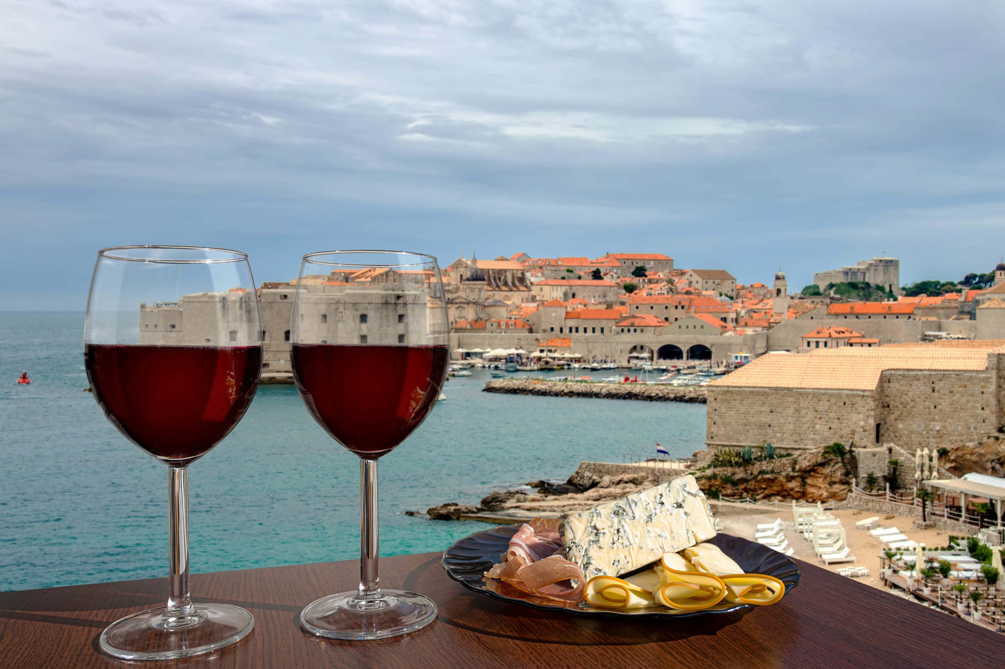 Two glasses of red wine and a plate of assorted cheeses and cured meats on a table overlooking the historic walled city and harbor of Dubrovnik, Croatia.