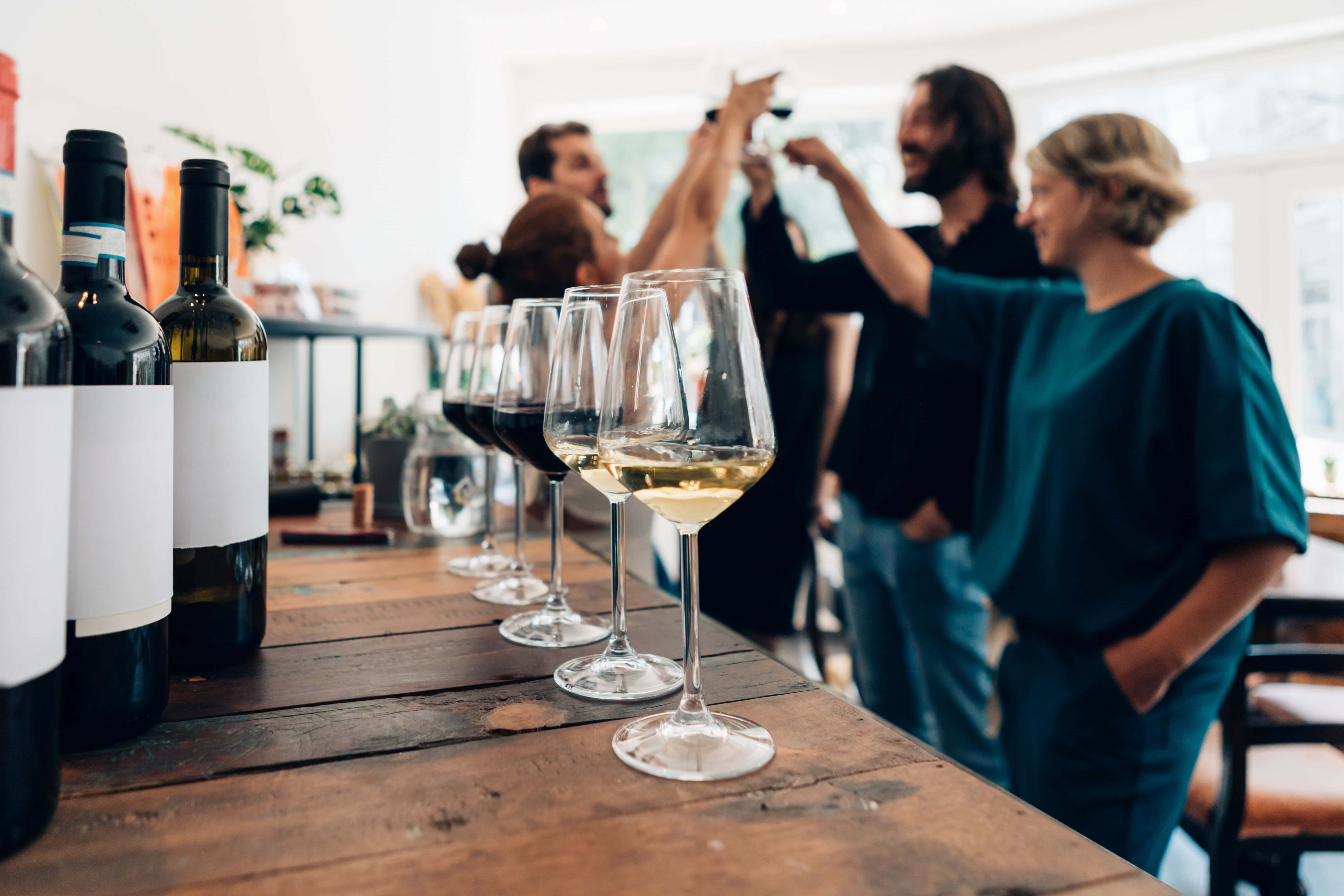 Close-up of wine glasses and bottles on a wooden table with a group of people in the background raising their glasses in a toast.