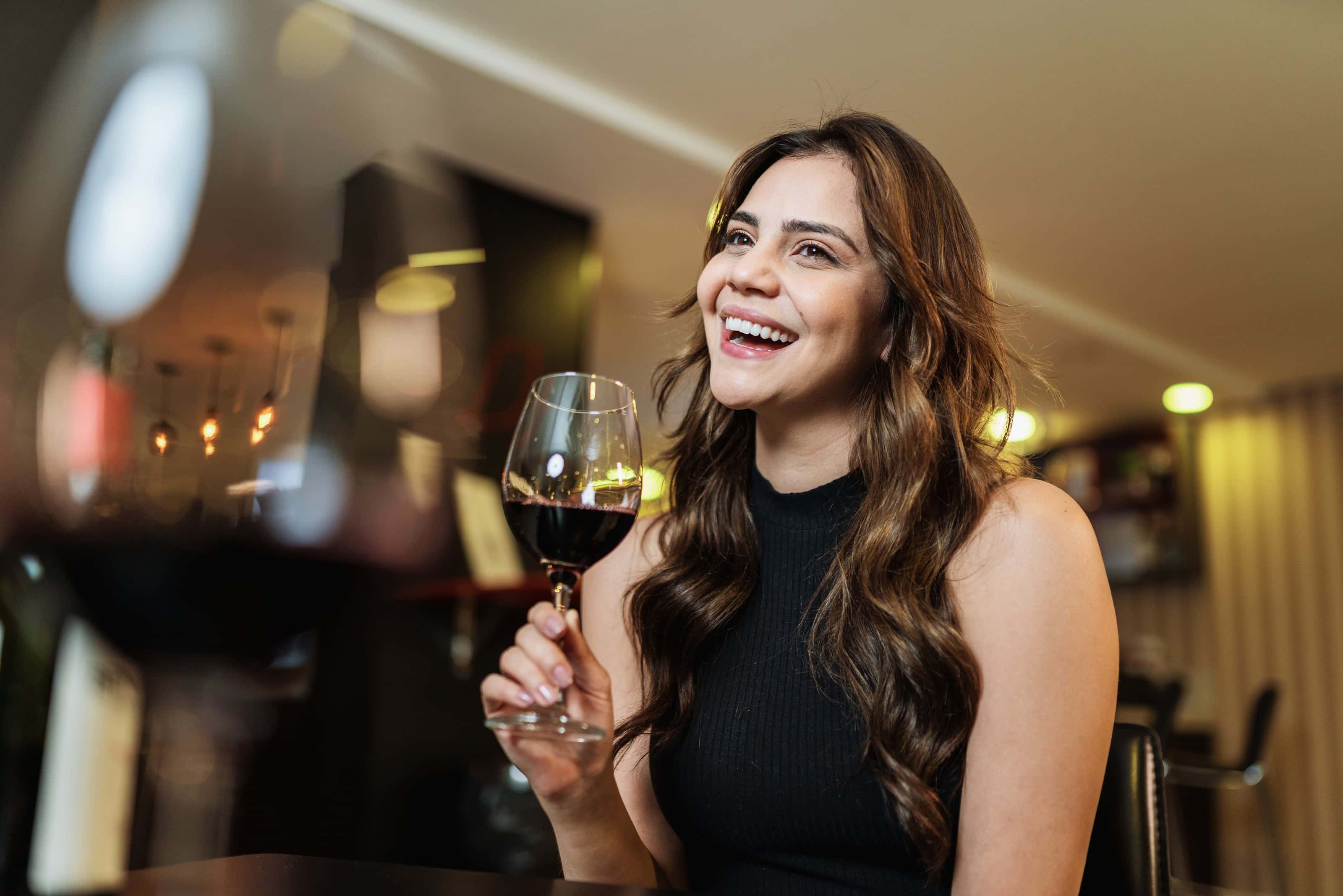 Smiling woman holding a glass of red wine in a warmly lit modern bar or restaurant, with blurred wine glasses and ambient lighting in the foreground.