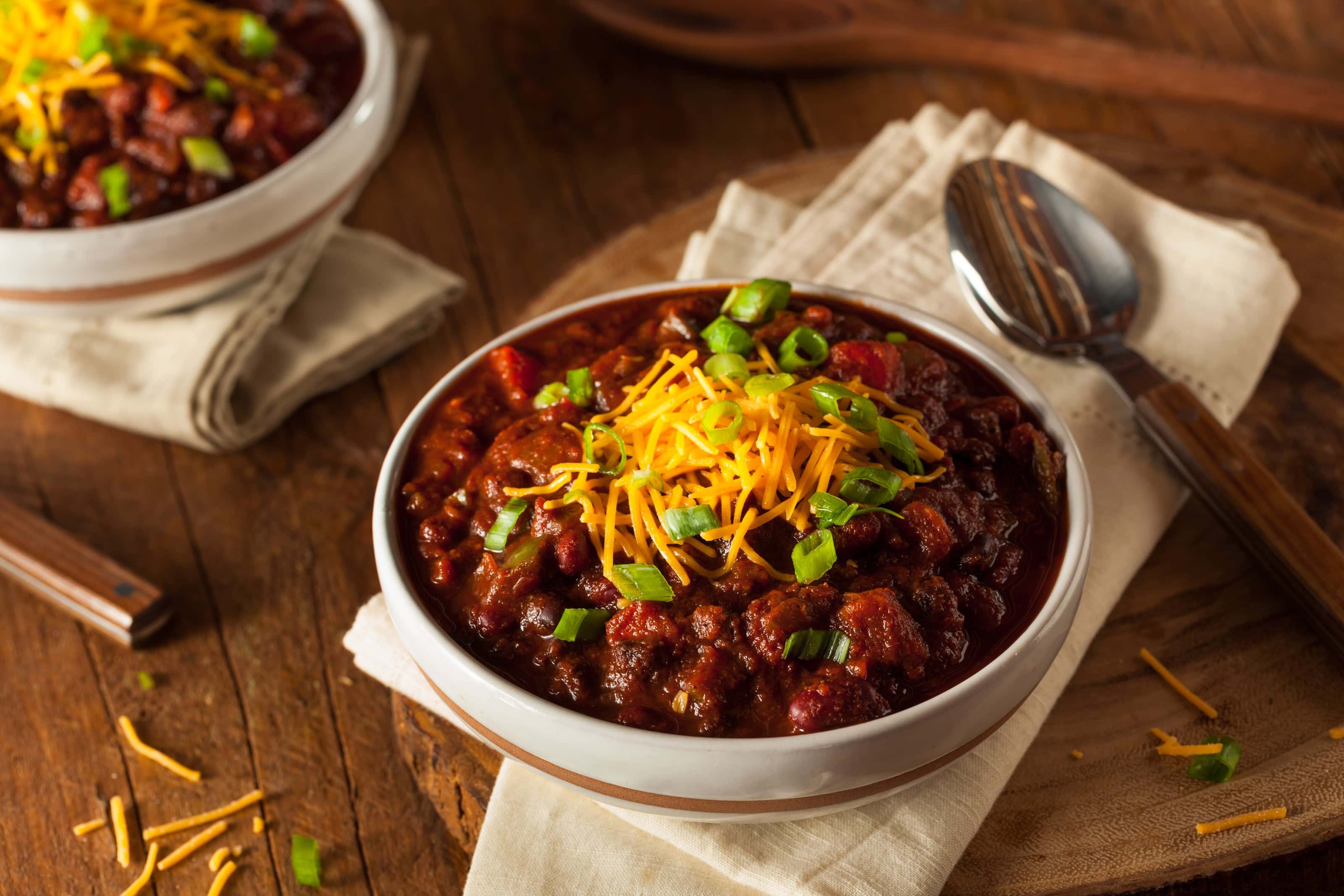 Bowl of hearty chili topped with shredded cheddar cheese and chopped green onions, served on a rustic wooden table with a spoon and napkin.