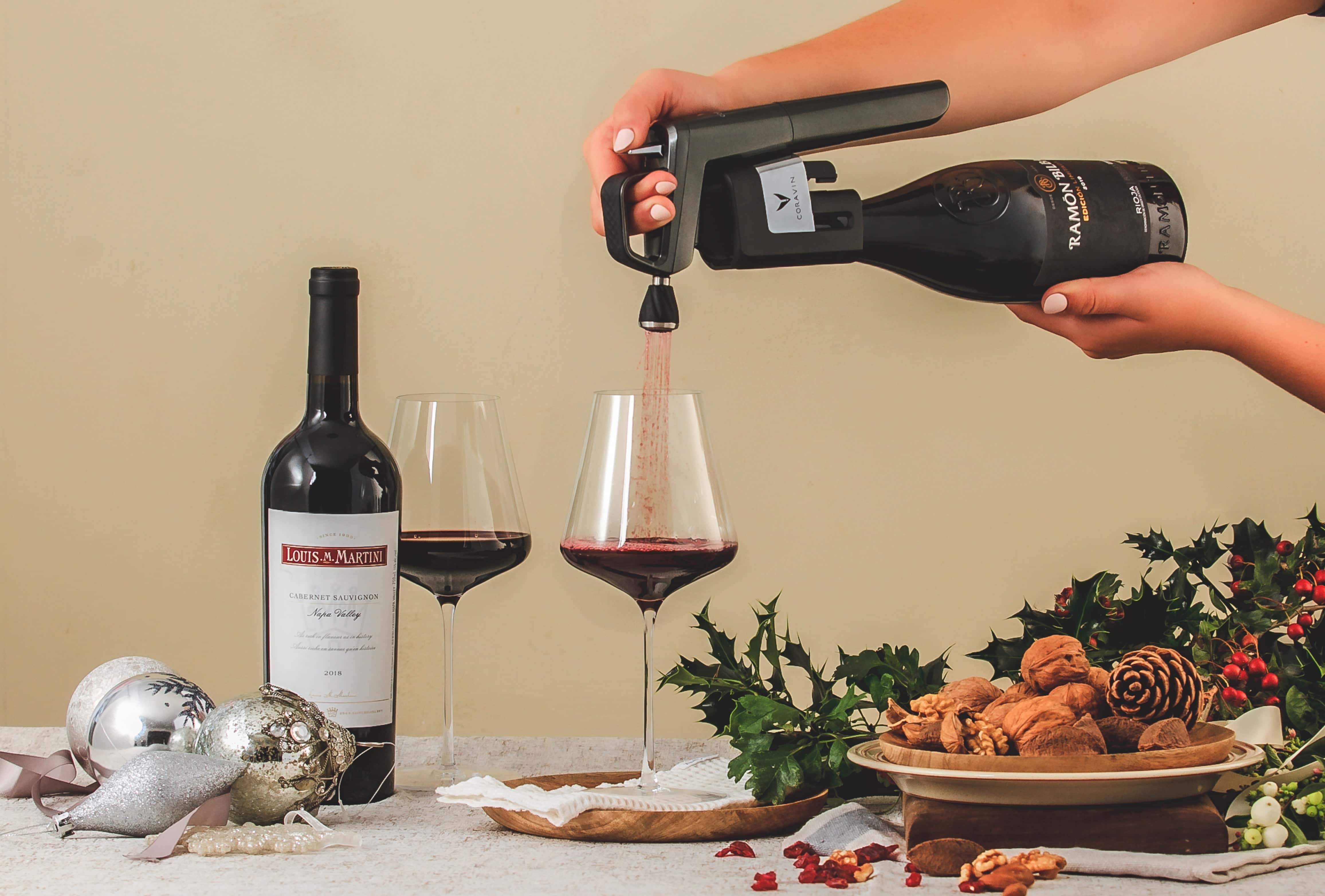 Person using a Coravin wine preservation system to pour red wine into a glass, with another glass and a Louis M. Martini wine bottle on the table, surrounded by festive holiday decor and a plate of nuts.