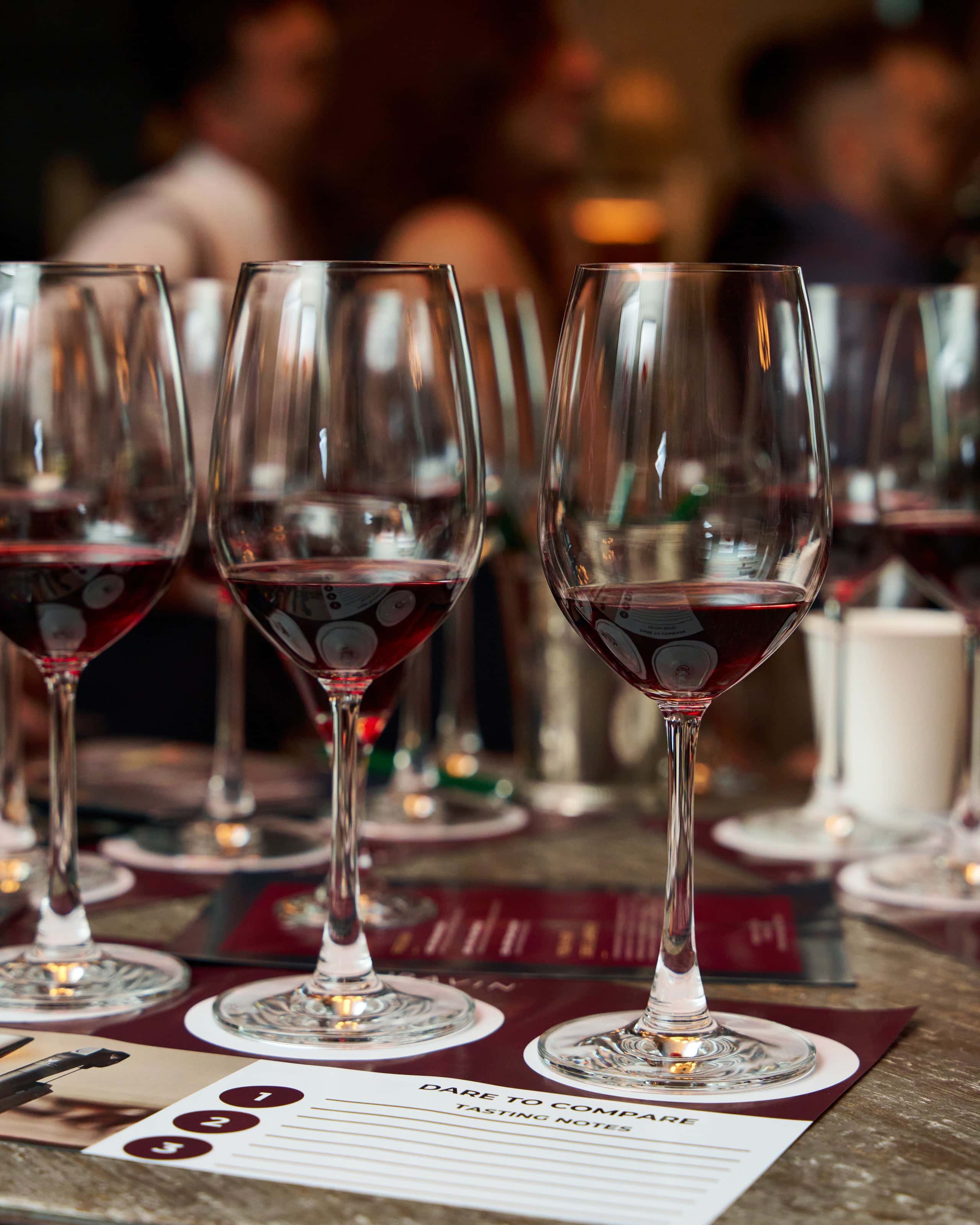 A row of wine glasses filled with red wine placed on tasting mats labeled 'Dare to Compare,' with blurred figures of people socializing in the background at a wine tasting event.