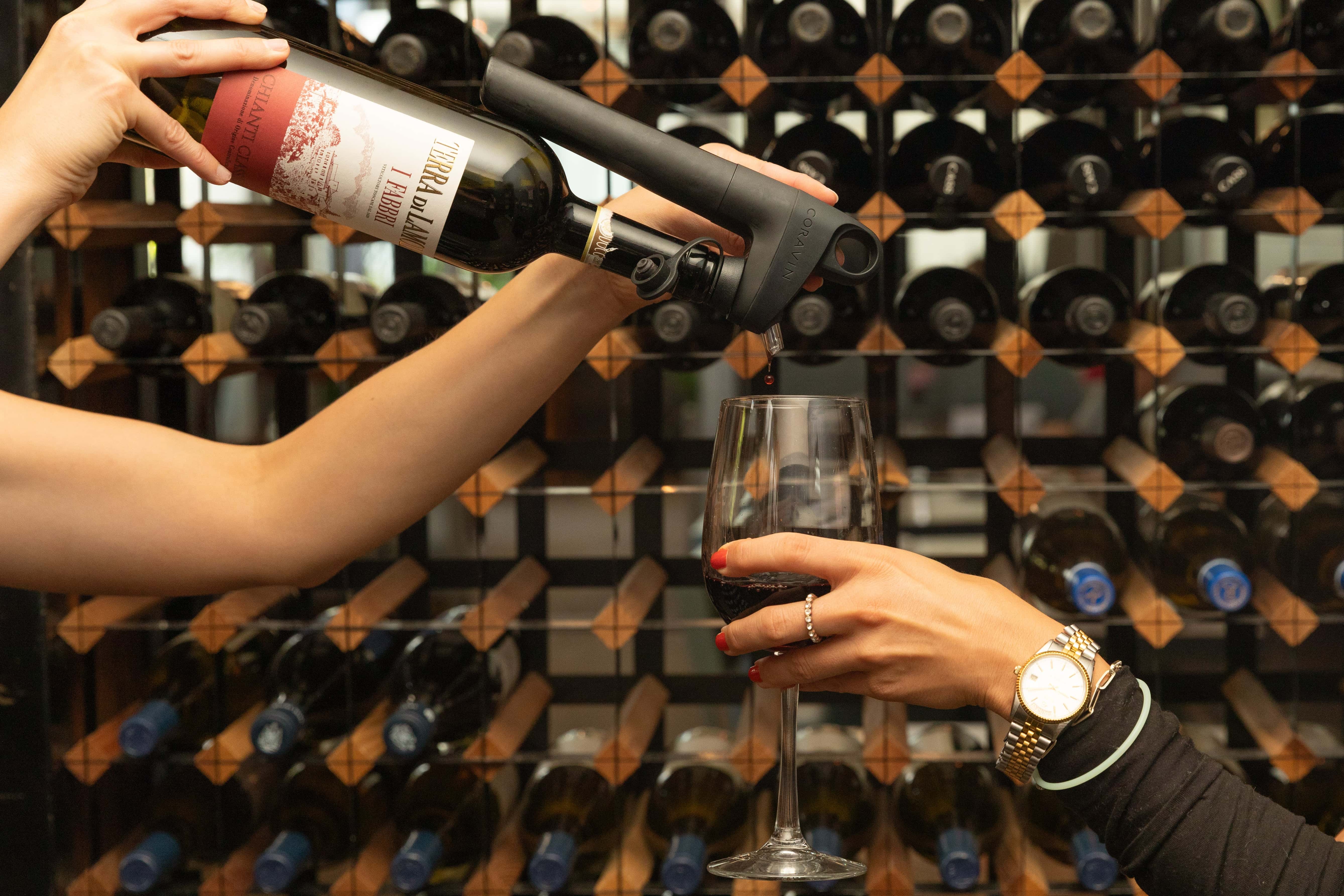 Using a Coravin wine preservation system to pour red wine into a glass in front of a wine rack filled with bottles.
