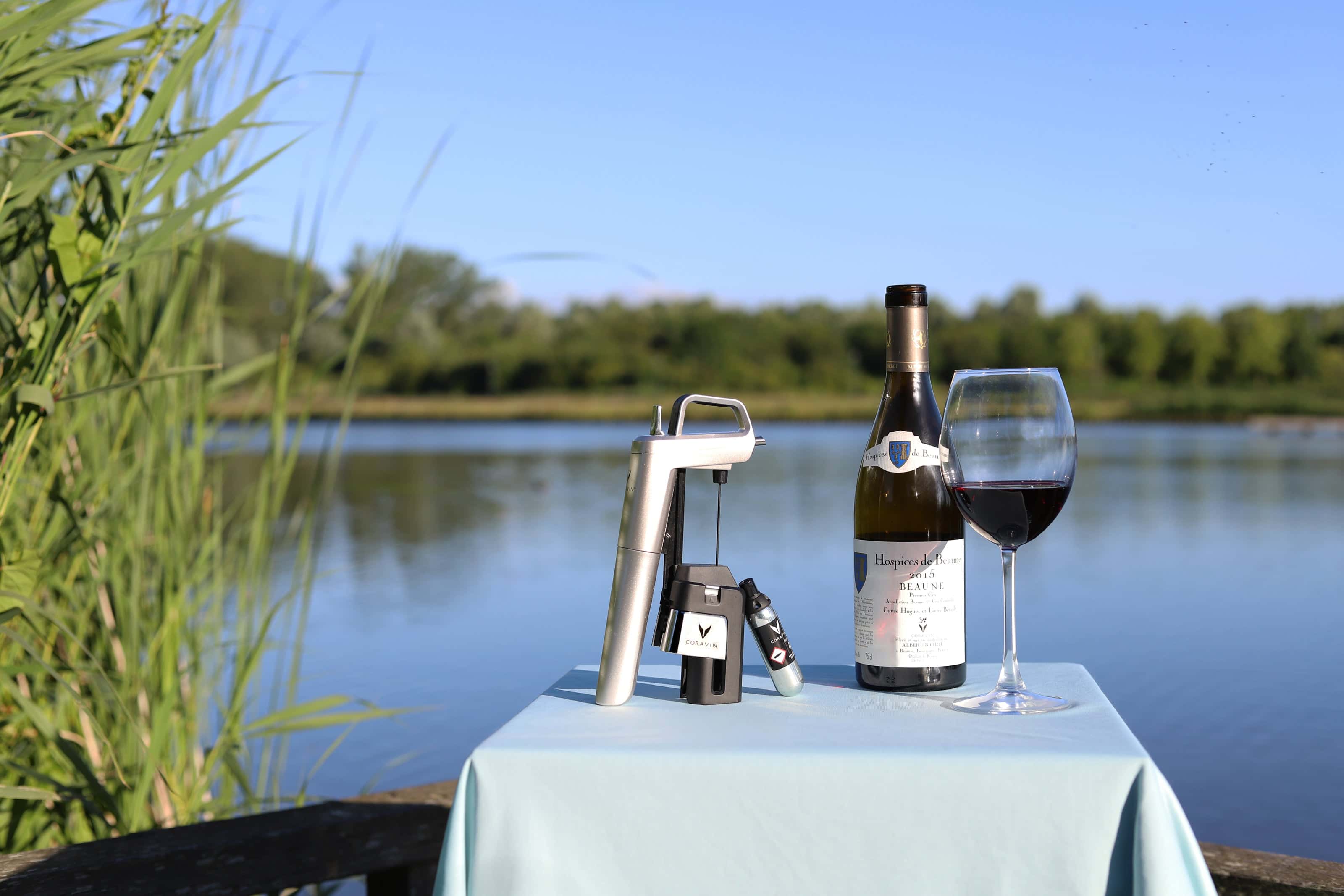 Coravin wine preservation system displayed outdoors on a table by a serene lake, alongside a bottle of Hospices de Beaune red wine and a filled wine glass, with tall grasses and a wooded shoreline in the background.