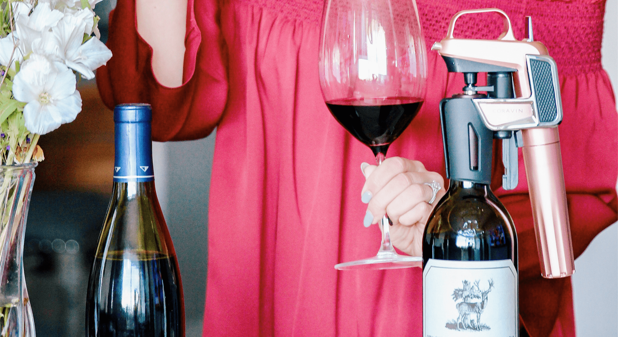 Woman in a red dress holding a glass of red wine next to two wine bottles, one equipped with a rose gold Coravin wine preservation system.