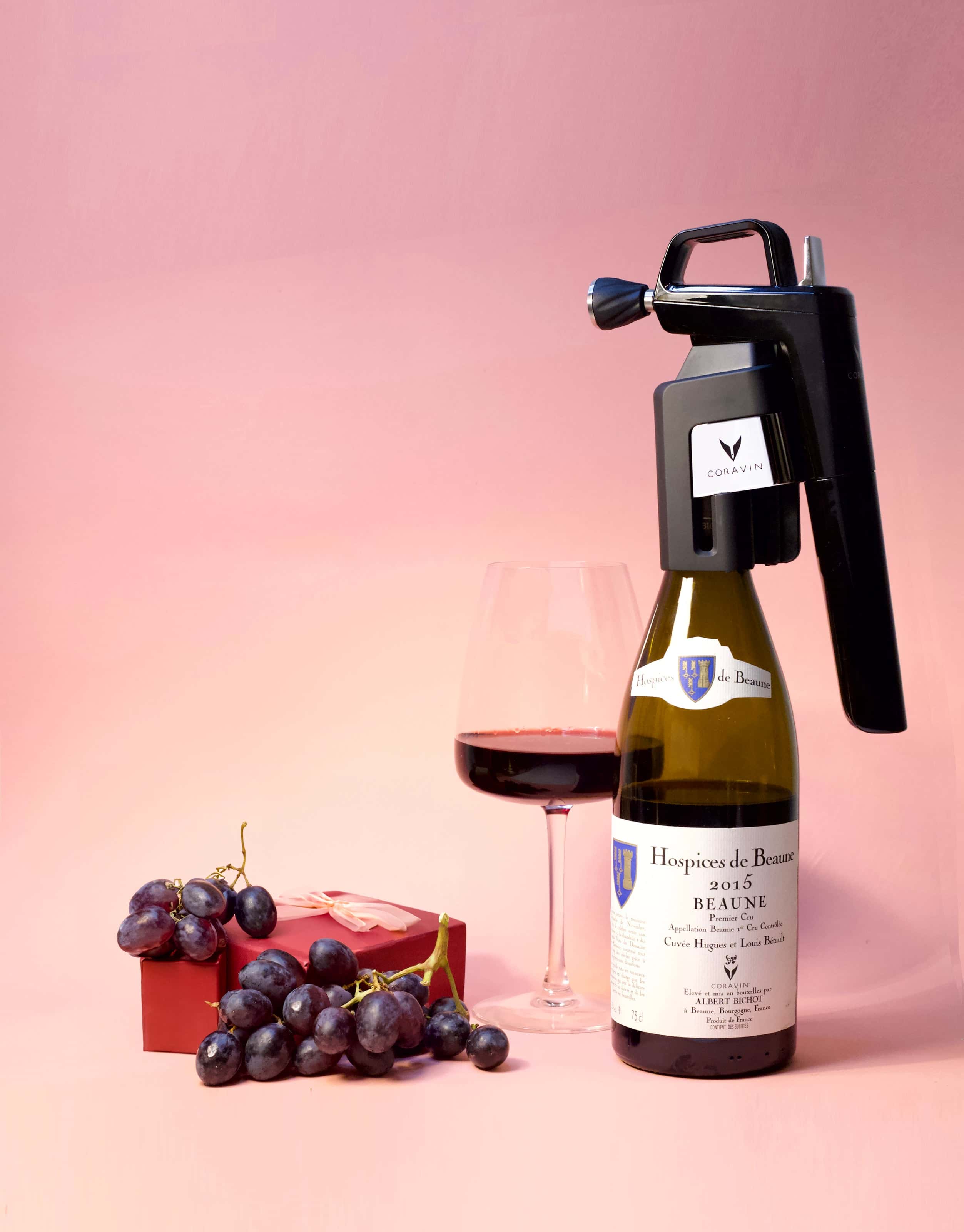 Coravin wine preservation system on a bottle of Hospices de Beaune 2015 red wine with a filled glass, fresh grapes, and a gift box on a pink background.