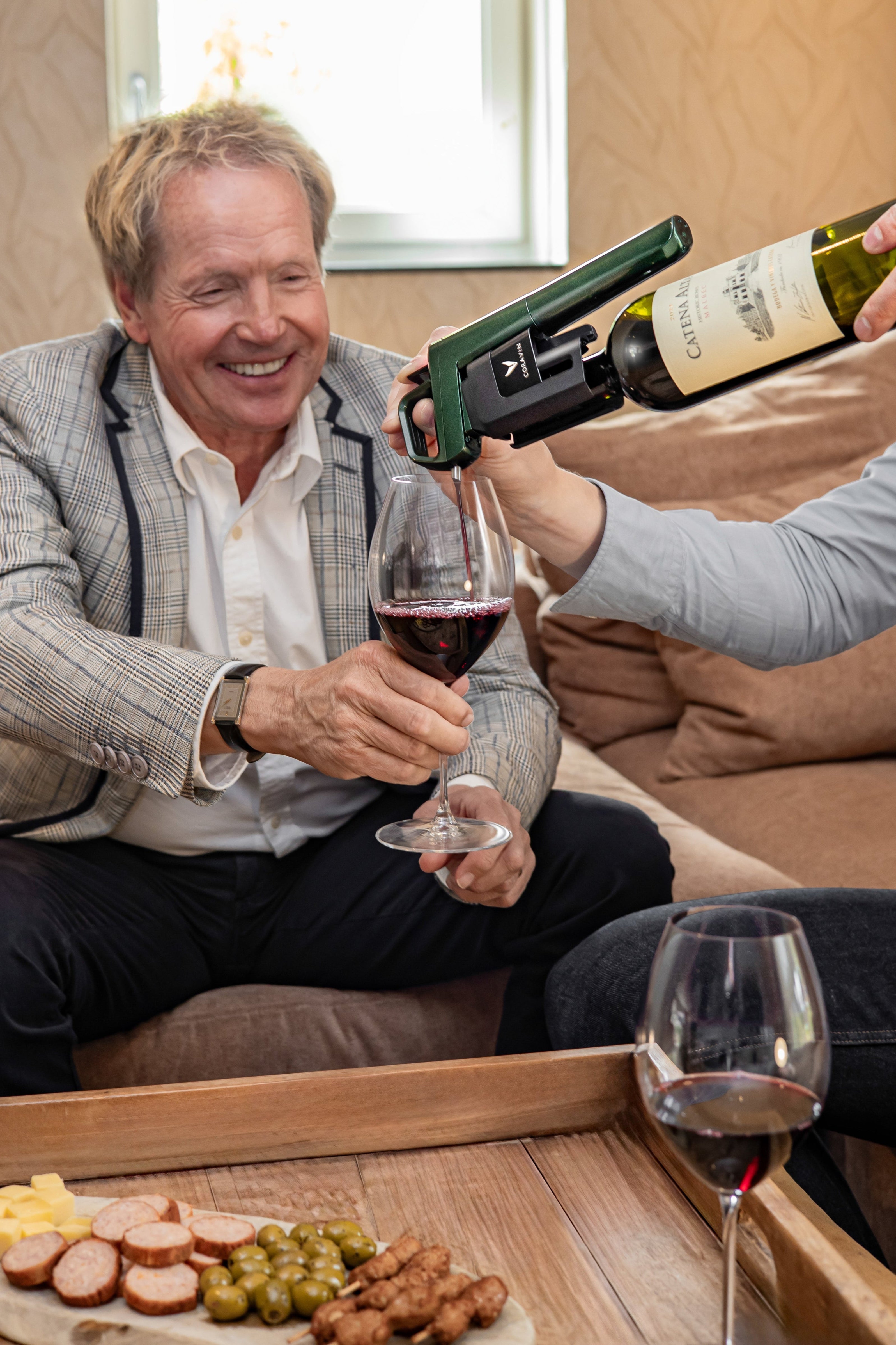 A man in a blazer smiles while a second person pours red wine into his glass, with snacks on a wooden tray in front.
